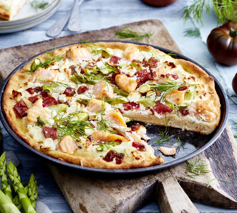 Angeschnittene Lachs-Feta-Pizza auf dunklem Teller. Drum herum Gemüse auf hellem Holzuntergrund.  | © EAT CLUB / Isabell Triemer