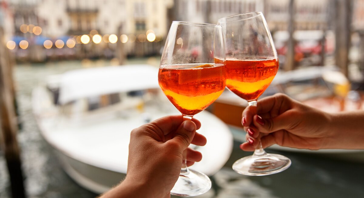 Zwei Hände stoßen mit zwei Gläsern Aperol Spritz an. Im Hintergrund sieht man unscharf die Kulisse eines Hafens. 