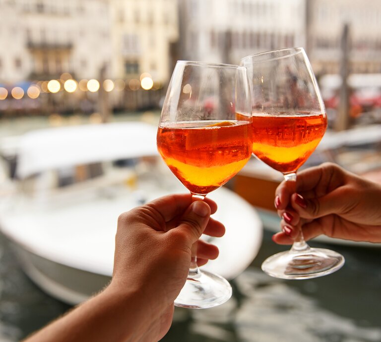 Zwei Hände stoßen mit zwei Gläsern Aperol Spritz an. Im Hintergrund sieht man unscharf die Kulisse eines Hafens.  | © Getty Images/kkshepel