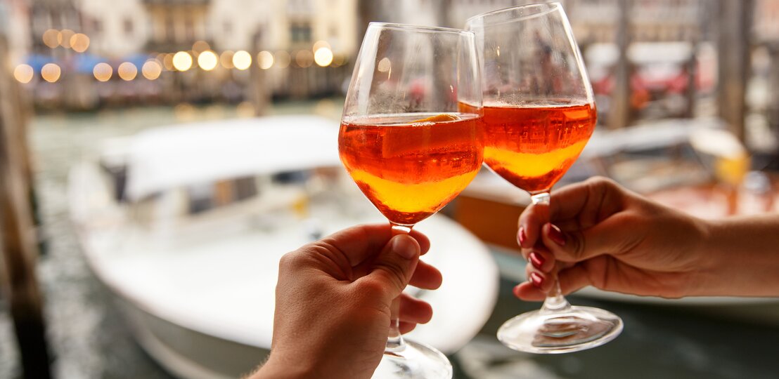 Zwei Hände stoßen mit zwei Gläsern Aperol Spritz an. Im Hintergrund sieht man unscharf die Kulisse eines Hafens.  | © Getty Images/kkshepel