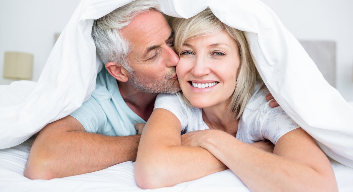 Älteres Ehepaar liegt im Bett mit der Bettdecke über den Köpfen, er küsst sie auf die Wange, sie lacht