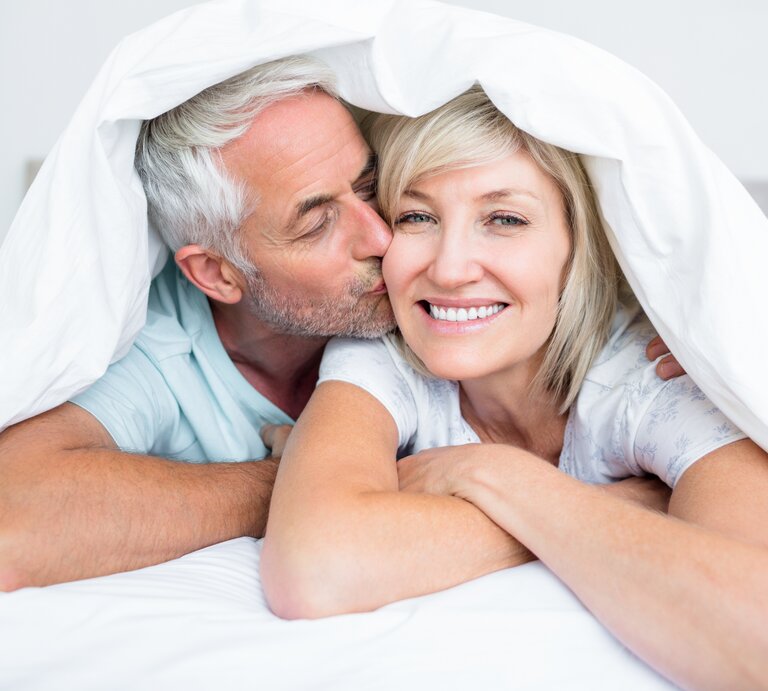 Älteres Ehepaar liegt im Bett mit der Bettdecke über den Köpfen, er küsst sie auf die Wange, sie lacht | © iStock.com/Wavebreakmedia
