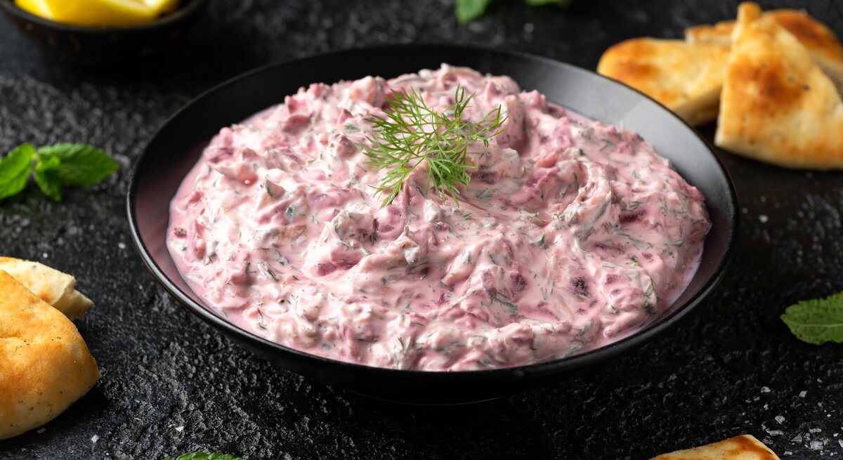 Auf einem dunklen Untergrund steht eine Schale mit Rote-Bete-Tsatsiki. Daneben liegen Kekse sowie mehrere Schnitzer Zitrone.