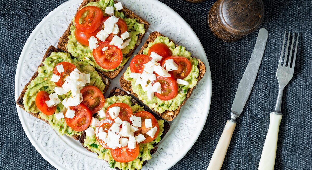 Vier Toasts auf einem weißen Teller belegt mit der Avocado-Basilikum-Creme, Tomate und Mozzarella. Der Untergrund ist eine graue Stoffdecke und neben dem Teller sind Besteck, Salz und Pfeffer. Alles direkt von oben fotografiert.