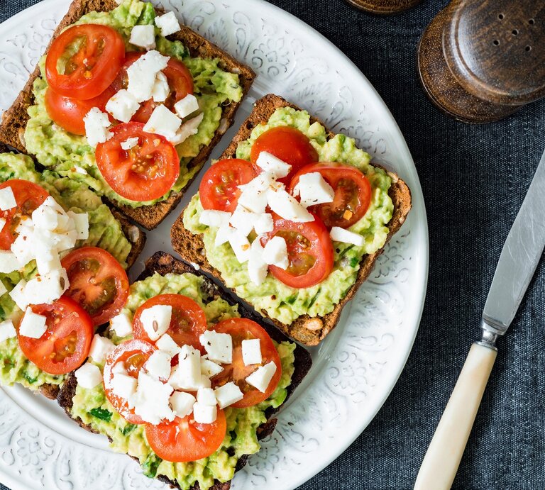 Vier Toasts auf einem weißen Teller belegt mit der Avocado-Basilikum-Creme, Tomate und Mozzarella. Der Untergrund ist eine graue Stoffdecke und neben dem Teller sind Besteck, Salz und Pfeffer. Alles direkt von oben fotografiert. | © Shutterstock/ Vladislav Noseek