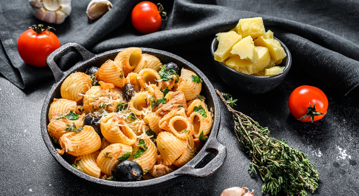 Draufsicht: eine runde schwarze Schüssel Pasta mit Oliven und Tomaten auf schwarzem Untergrund. Drum herum liegen frische Tomaten, frischer Thymian, frischer Knoblaucg und eine Schale mit Parmesanstücken.