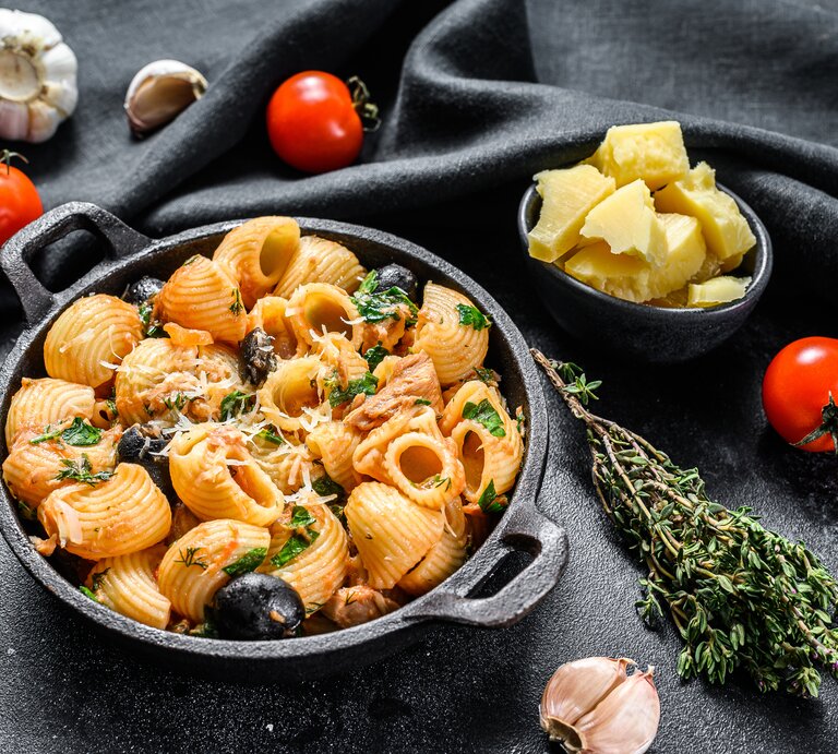 Draufsicht: eine runde schwarze Schüssel Pasta mit Oliven und Tomaten auf schwarzem Untergrund. Drum herum liegen frische Tomaten, frischer Thymian, frischer Knoblaucg und eine Schale mit Parmesanstücken. | © Shutterstock / Mironov Vladimir