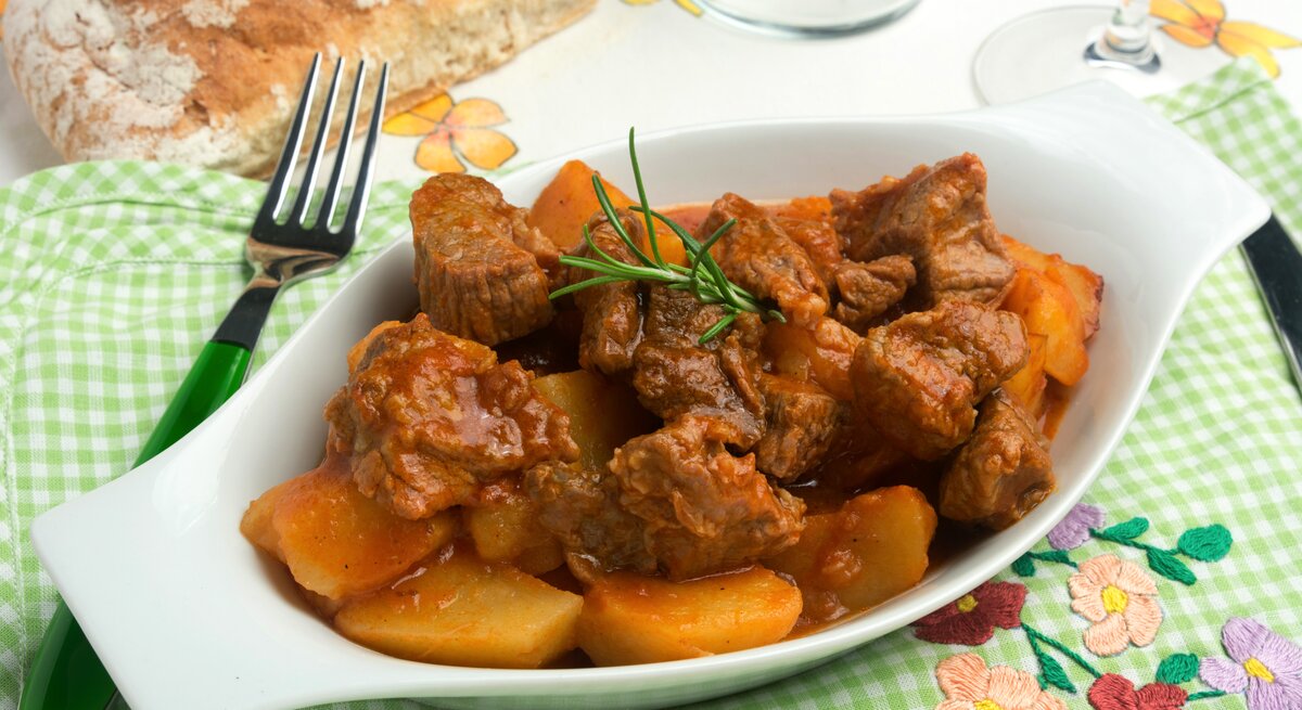 Draufsicht schräg: Auf einem grün-weiß-kariertem Tuch ist eine kleine Schale mit Spezzatino di manzo, ein italienisches Gulasch. Drum herum liegen eine Gabel und ein Stück Weißbrot.