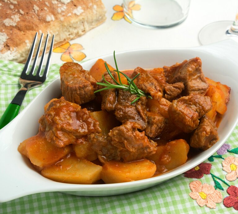 Draufsicht schräg: Auf einem grün-weiß-kariertem Tuch ist eine kleine Schale mit Spezzatino di manzo, ein italienisches Gulasch. Drum herum liegen eine Gabel und ein Stück Weißbrot. | © Shutterstock / Elflaco1983