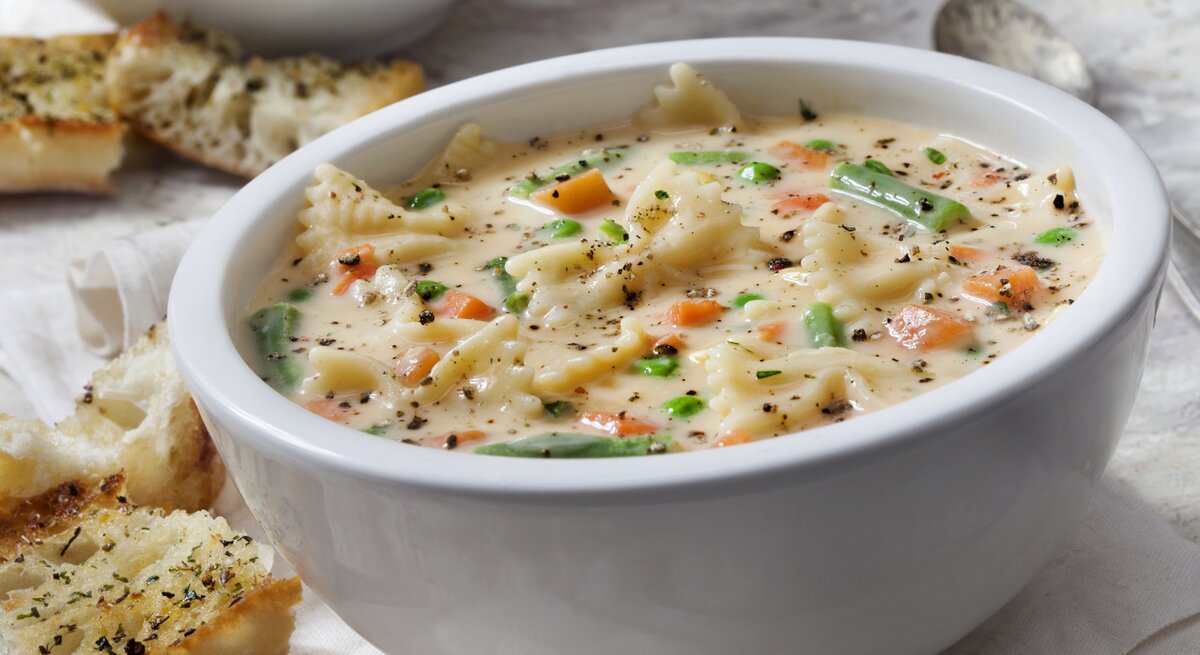 Suppenteller gefüllt mit Käse-Nudelsuppe mit Gemüse auf hellem Untergrund.