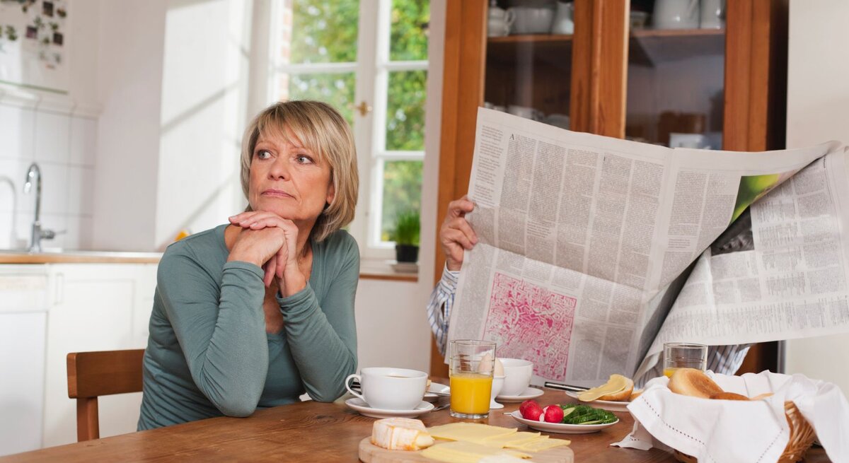 Eine blonde Frau mittleren Alters sitzt nachdenklich am Frühstückstisch, während ein Mann hinter einer Zeitung verborgen ist.