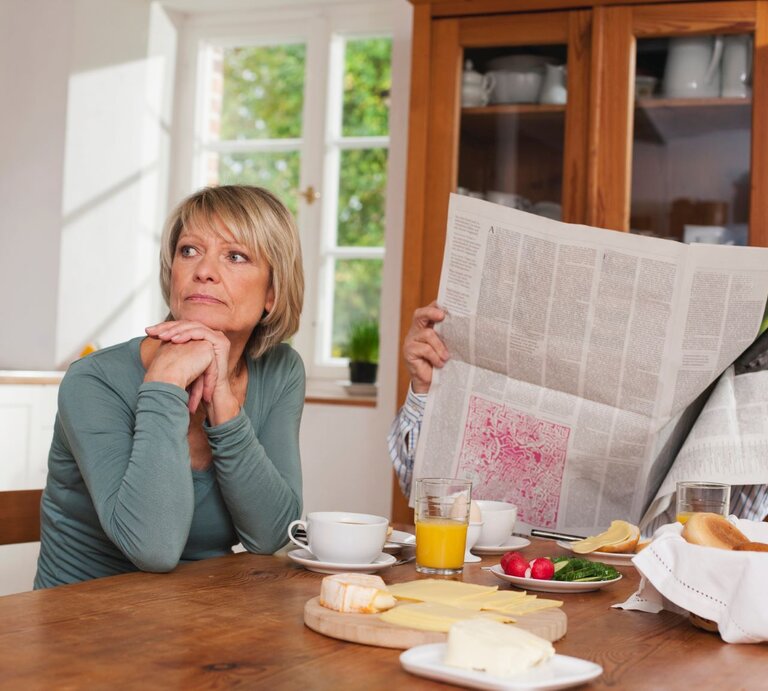 Eine blonde Frau mittleren Alters sitzt nachdenklich am Frühstückstisch, während ein Mann hinter einer Zeitung verborgen ist. | © Getty Images / Westend61