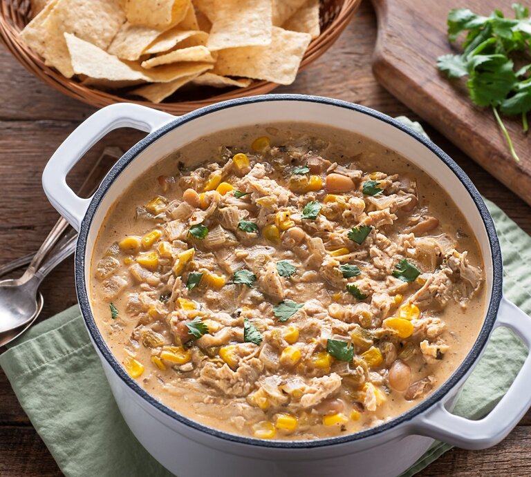 Ein weißer Topf mit dem weißen Chili con Carne auf einem dunklen Holztisch. Oben rechts im Bild grüne Chili, Koriander und eine Schale mit Maischips. | © GettyImages/ rudisill