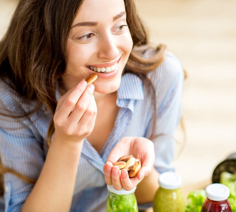 Eine junge Frau mit langen braunen Haaren und einem blau gestreiften Hemd sitzt lächelnd an einem Tisch und isst Nüsse aus ihrer Handfläche. Vor ihr stehen frische grüne Smoothies und ein Salat, was auf eine gesunde Ernährung hindeutet. Sie scheint eine Pause zu genießen und wirkt entspannt und zufrieden in ihrer hellen, freundlichen Umgebung. | © iStock.com/RossHelen