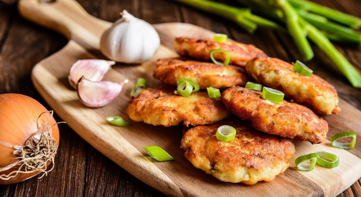 Mehrere Thunfischbratlinge auf einem Holzbrett und einem rustikalen Holztisch mit Knoblauch und Frühlingszwiebel im Hintergrund.