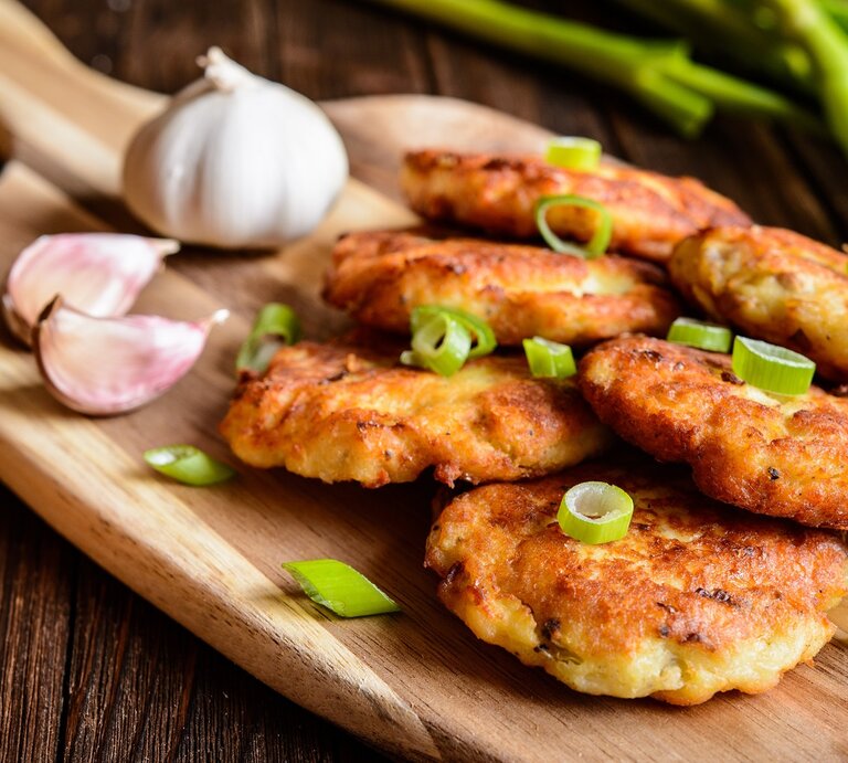 Mehrere Thunfischbratlinge auf einem Holzbrett und einem rustikalen Holztisch mit Knoblauch und Frühlingszwiebel im Hintergrund. | © Shutterstock/NoirChocolate