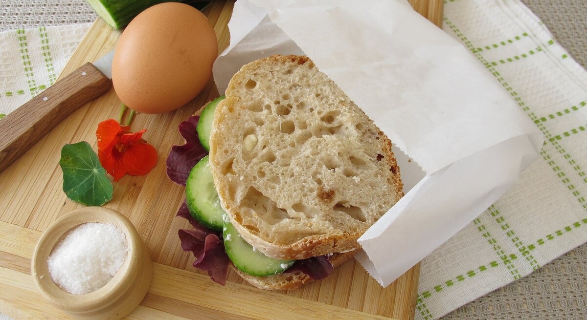 Ein belegtes Brot mit Butterbrottüte, ein Ei, eine Gurke, ein Messer und ein Schälchen Salz liegen auf einem Küchenbrett aus Holz.