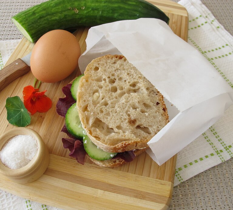 Ein belegtes Brot mit Butterbrottüte, ein Ei, eine Gurke, ein Messer und ein Schälchen Salz liegen auf einem Küchenbrett aus Holz. | © Adobe Stock/Heike Rau