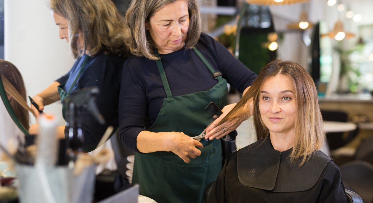 Eine ältere Friseurin mit grauen Haaren schneidet ihrer jüngeren Kundin, die blonde Haare hat und auf dem Friseurstuhl sitzt, die Haare. Vielleicht bekommt Sie eine der Trendfrisuren im Herbst 2023 geschnitten.