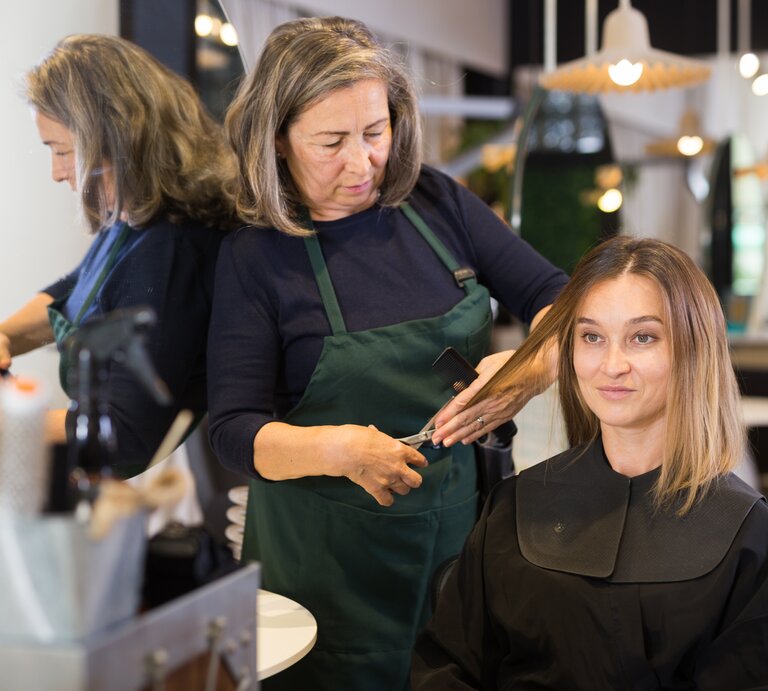Eine ältere Friseurin mit grauen Haaren schneidet ihrer jüngeren Kundin, die blonde Haare hat und auf dem Friseurstuhl sitzt, die Haare. Vielleicht bekommt Sie eine der Trendfrisuren im Herbst 2023 geschnitten. | © Shutterstock/ BearFotos