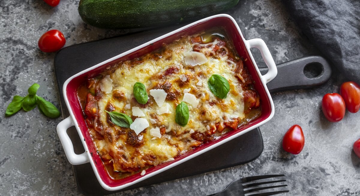 Form mit Lasagne auf grauem Untergrund. Drumherum liegen Tomaten, Zucchini und Basilikum-Blättchen.  | © Getty Images/Westend61