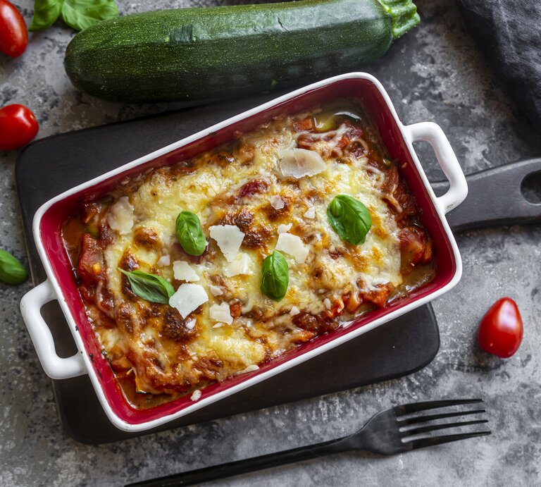 Form mit Lasagne auf grauem Untergrund. Drumherum liegen Tomaten, Zucchini und Basilikum-Blättchen.  | © Getty Images/Westend61