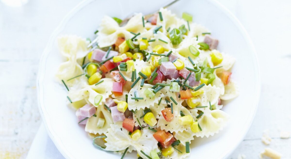 In einer Schüssel ist eine Portion Nudelsalat mit Farfalle, Schinkenwürfeln und Mais angerichtet.