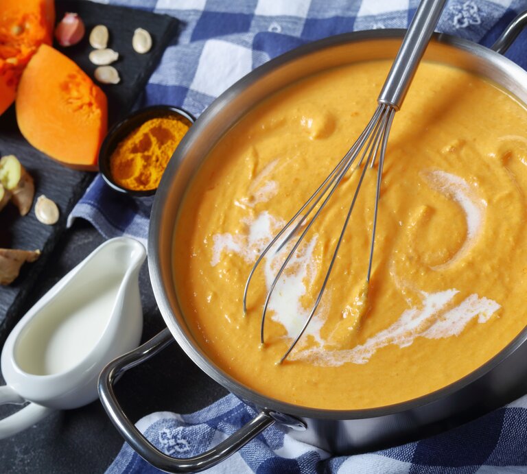 Ein Topf mit der Kürbissuppe auf einem Tisch mit blau-weiß-karierter Tischdecke.  | © iStock.com/from_my_point_of_view
