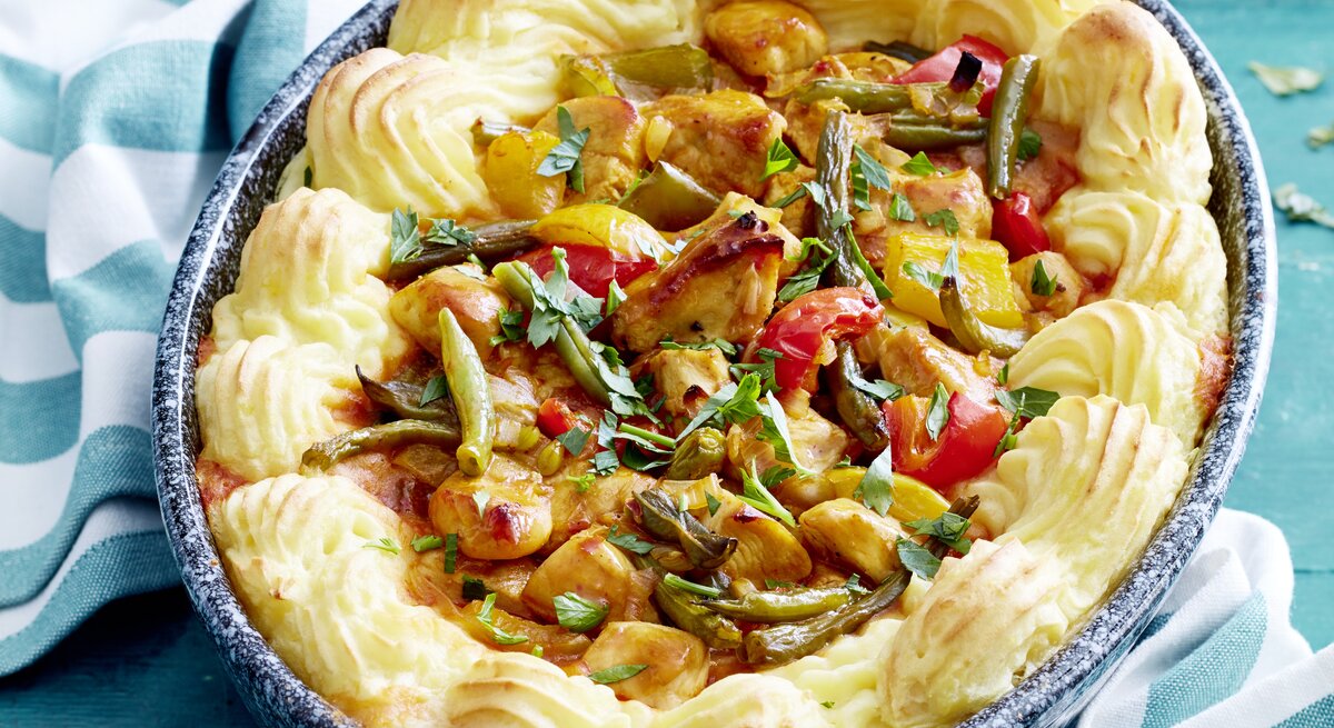 Eine Auflaufform mit Hähnchengulasch mit Kartoffelrand auf einem blau gestreiften Küchentuch.