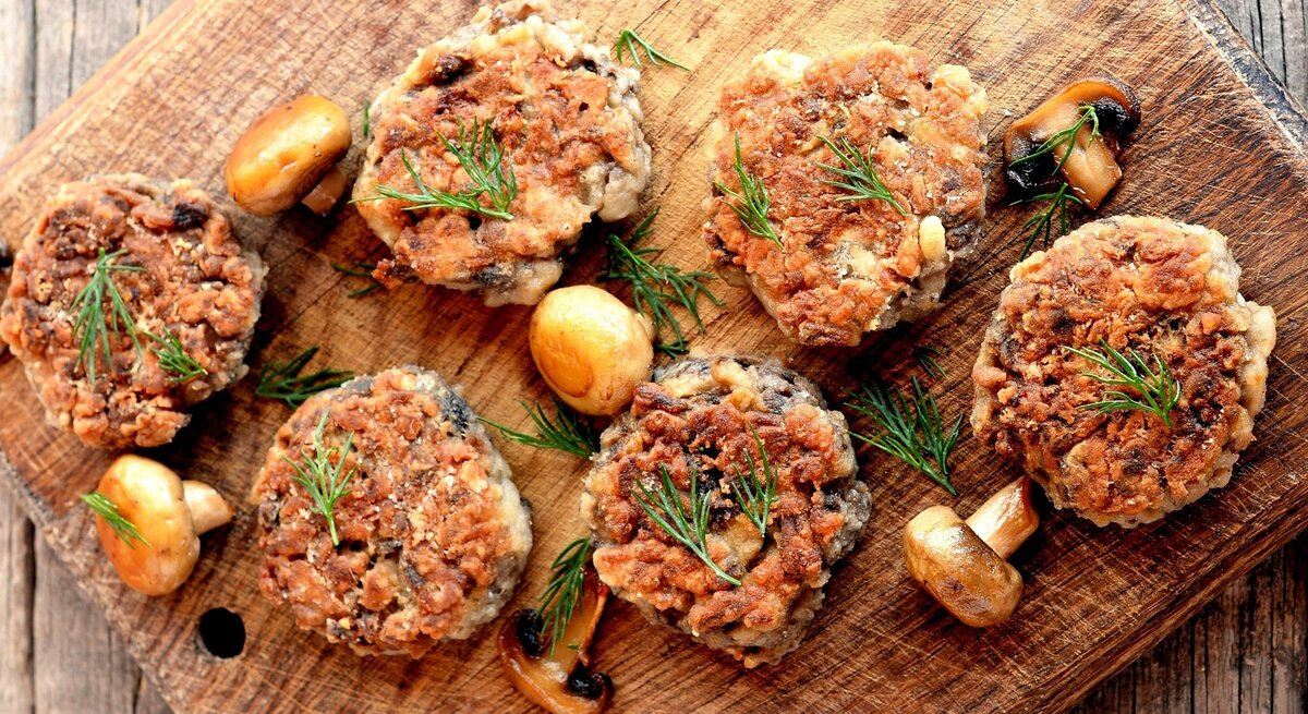 Mehrere Champignon-Bouletten auf einem Holzbrett mit gebratenen Pilzen und Kräutern.