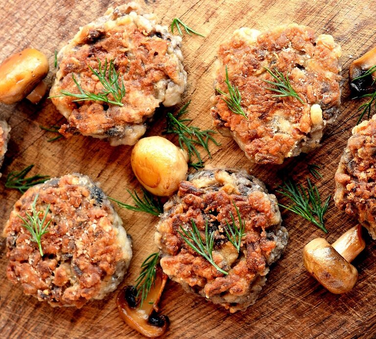 Mehrere Champignon-Bouletten auf einem Holzbrett mit gebratenen Pilzen und Kräutern. | © Shutterstock/ OnlyZoia