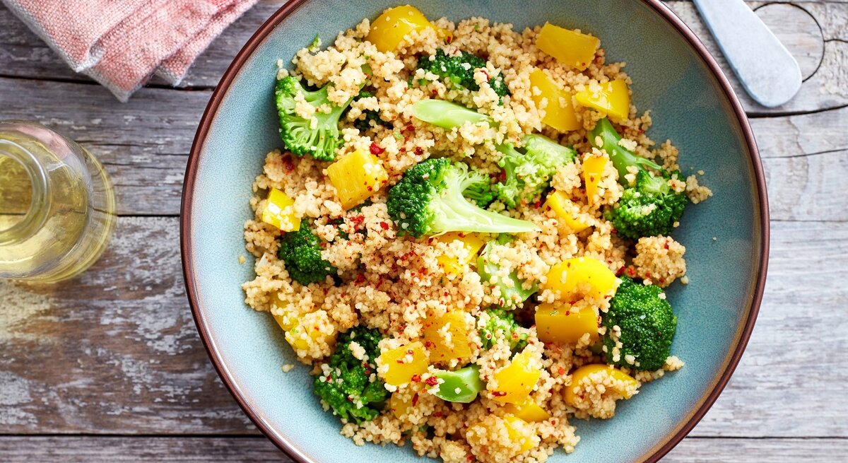 Brokkoli-Bowl mit Couscous in blauer Schüssel auf Holzuntergrund. Daneben rotes Küchentuch, Glasbehälter und das Ende einer Gabel/eines Löffels. 