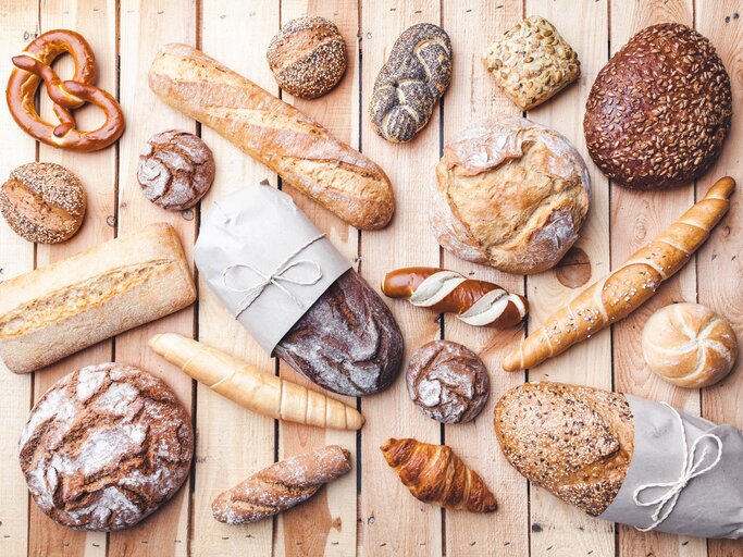 Mehrere Brote und Gebäck auf einem hellen Holztisch. Alles direkt von oben fotografiert. | © iStock/AND-ONE