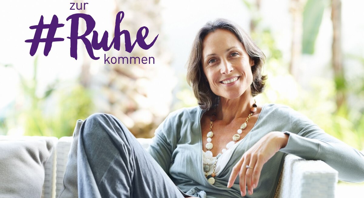 Eine lächelnde Frau mit mittellangen dunklen Haaren entspannt sich in einem hellen, gemütlichen Sessel. Sie trägt ein lässiges, graues Outfit und eine große, auffällige Halskette. Ihr entspanntes Auftreten und ihr zufriedenes Lächeln vermitteln ein Gefühl von Gelassenheit und Zufriedenheit. Im Hintergrund ist eine natürliche, grüne Umgebung unscharf zu sehen. Über ihr steht in großer lila Schrift der Hashtag "#Ruhe kommen", was darauf hinweist, dass sie sich in einem Moment der Ruhe und des Wohlbefindens befindet.