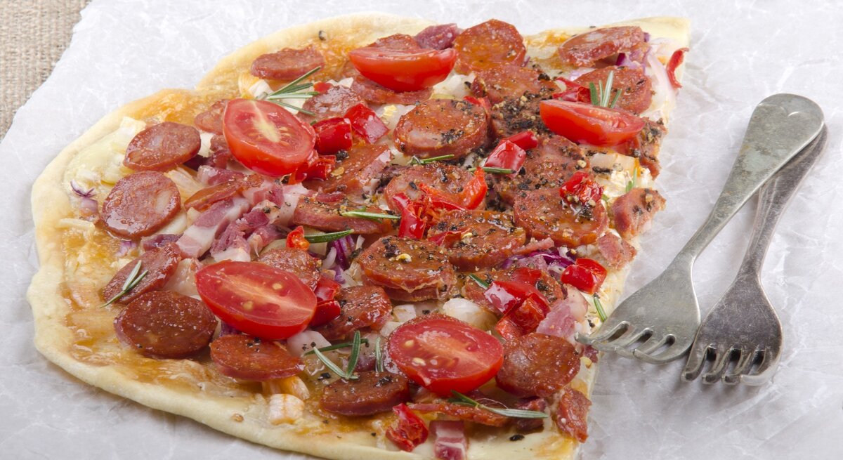Die Hälfte vom Flammkuchen auf Backpapier mit zwei Gabeln auf einer hellen Tischdecke.