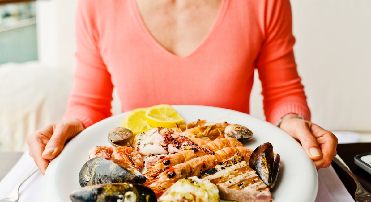Eine Person im rosa Oberteil hält einen Teller mit Garnelen, Muscheln, Tintenfisch und Zitronenscheiben.