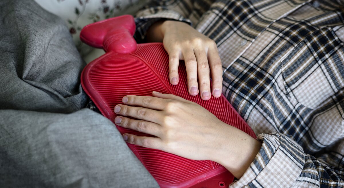 Das Bild zeigt eine Person, die entspannt auf einer Couch oder einem Bett liegt, mit einer rot gefärbten Wärmflasche auf ihrem Bauch. Die Person trägt eine karierte Jacke und liegt unter einer grauen Decke. Ihre Hände liegen sanft auf der Wärmflasche, was darauf hindeuten könnte, dass sie Wärme und Komfort sucht, möglicherweise um Schmerzen oder Unbehagen zu lindern. Das Muster der Jacke und die Textur der Wärmflasche sind deutlich sichtbar und erzeugen einen gemütlichen und behaglichen Eindruck.