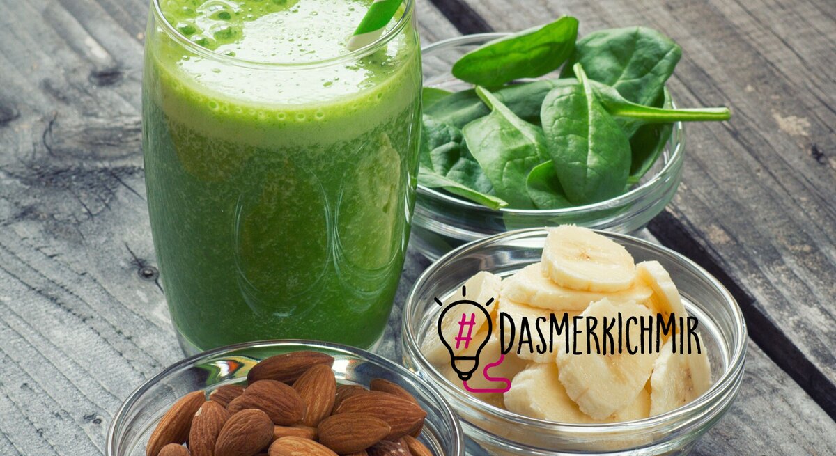 Ein frisch zubereiteter grüner Smoothie in einem Glas mit einem gestreiften Strohhalm wird präsentiert neben Schalen mit gesunden Zutaten: frische Spinatblätter und geschnittene Bananenstücke. Eine weitere Schale enthält rohe Mandeln. Diese Anordnung von nährstoffreichen Lebensmitteln auf einer rustikalen Holzoberfläche symbolisiert gesunde Ernährung und Brainfood, was durch den Hashtag "#DAS MERK ICH MIR" neben einer Glühbirnen-Grafik verstärkt wird.