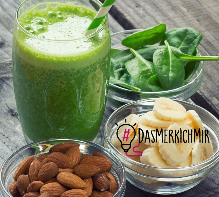 Ein frisch zubereiteter grüner Smoothie in einem Glas mit einem gestreiften Strohhalm wird präsentiert neben Schalen mit gesunden Zutaten: frische Spinatblätter und geschnittene Bananenstücke. Eine weitere Schale enthält rohe Mandeln. Diese Anordnung von nährstoffreichen Lebensmitteln auf einer rustikalen Holzoberfläche symbolisiert gesunde Ernährung und Brainfood, was durch den Hashtag "#DAS MERK ICH MIR" neben einer Glühbirnen-Grafik verstärkt wird. | © iStock.com/Lecic
