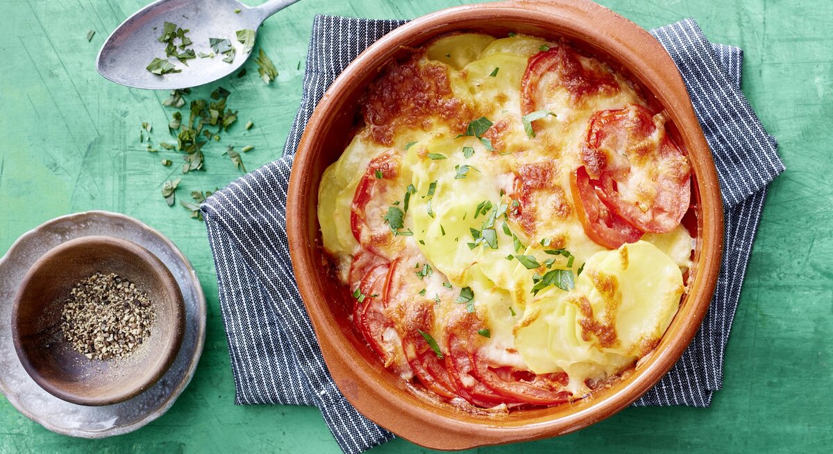 Tomaten-Kartoffel-Auflauf, überbacklen mit Käse und bestreut mit frischen Kräutern. Serviert in einer großen, runden Auflaufform auf einem türkisenen Untergrund. 