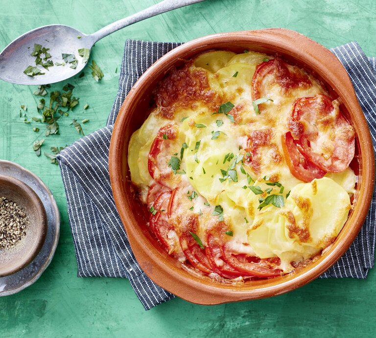Tomaten-Kartoffel-Auflauf, überbacklen mit Käse und bestreut mit frischen Kräutern. Serviert in einer großen, runden Auflaufform auf einem türkisenen Untergrund.  | © EAT CLUB / Isabell Triemer