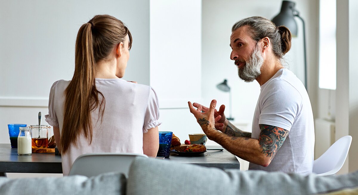 Paar sitzt auf einer Couch beim Frühstück und unterhält sich gestikulierend 