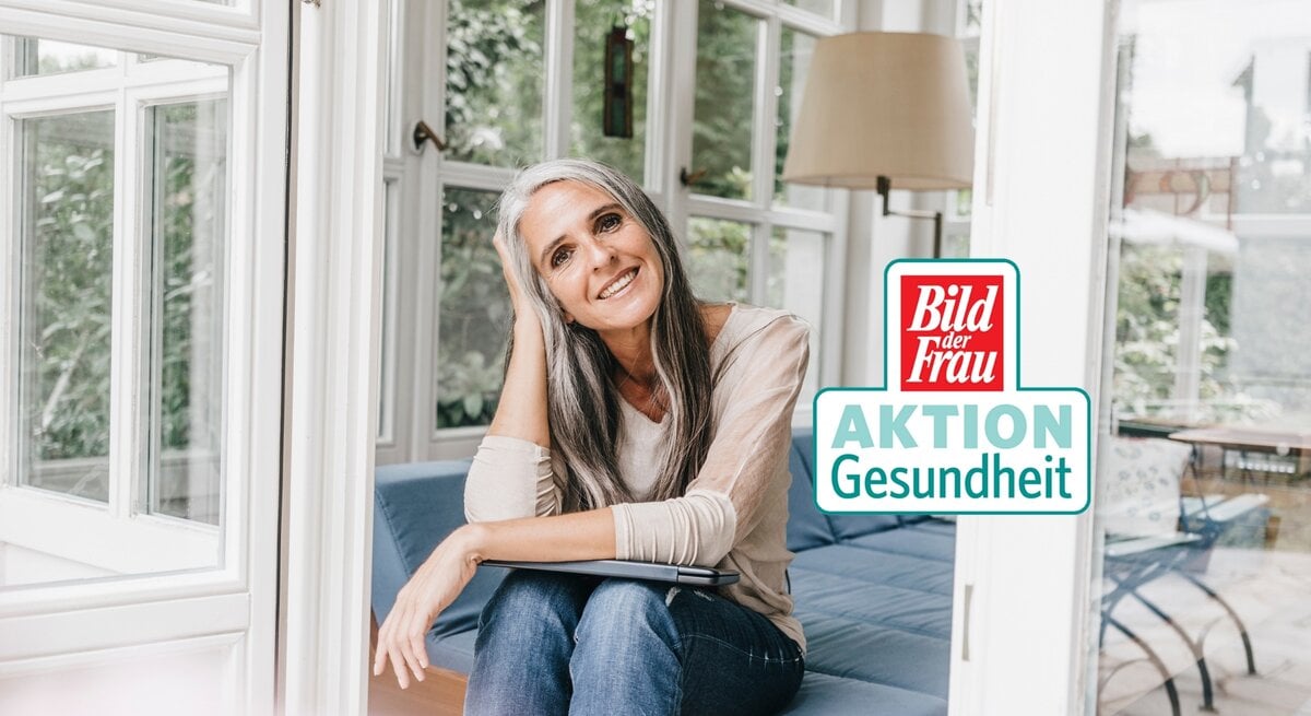Eine Frau mittleren Alters mit graumelierten Haaren und einem warmen Lächeln sitzt entspannt auf einer blauen Couch vor einem großen Fenster. Sie trägt ein lässiges, beiges Langarmshirt und dunkelblaue Jeans. Das Zimmer ist hell und freundlich, mit einem Blick auf grüne Pflanzen außerhalb des Fensters. Auf dem Bild ist auch ein Logo "Bild der Frau AKTION Gesundheit" erkennbar.