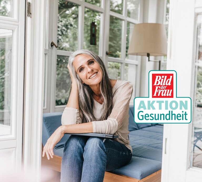 Eine Frau mittleren Alters mit graumelierten Haaren und einem warmen Lächeln sitzt entspannt auf einer blauen Couch vor einem großen Fenster. Sie trägt ein lässiges, beiges Langarmshirt und dunkelblaue Jeans. Das Zimmer ist hell und freundlich, mit einem Blick auf grüne Pflanzen außerhalb des Fensters. Auf dem Bild ist auch ein Logo "Bild der Frau AKTION Gesundheit" erkennbar. | © GettyImages/Westend61 [M]
