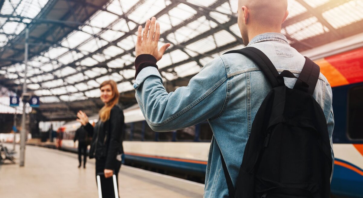 Zwei Personen verabschieden sich an einem Bahnhof voneinander. Im Vordergrund steht eine Person mit dem Rücken zur Kamera, die eine Jeansjacke und einen schwarzen Rucksack trägt und der anderen Person zuwinkt. Die zweite Person steht weiter hinten auf dem Bahnsteig, ebenfalls winkend, und hält einen Koffer. Im Hintergrund ist ein Zug zu sehen.