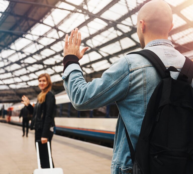 Zwei Personen verabschieden sich an einem Bahnhof voneinander. Im Vordergrund steht eine Person mit dem Rücken zur Kamera, die eine Jeansjacke und einen schwarzen Rucksack trägt und der anderen Person zuwinkt. Die zweite Person steht weiter hinten auf dem Bahnsteig, ebenfalls winkend, und hält einen Koffer. Im Hintergrund ist ein Zug zu sehen. | © iStock.com/Circle Creative Studio