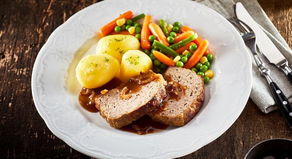 Draufsicht: Auf einem rustikalen Holztisch steht ein weißer Teller mit Omas Hackbraten mit Soße und Erbsen-Möhren-Bohnen-Gemüse. Daneben liegen eine Serviette aus grobem Leinen und ein Besteck.
