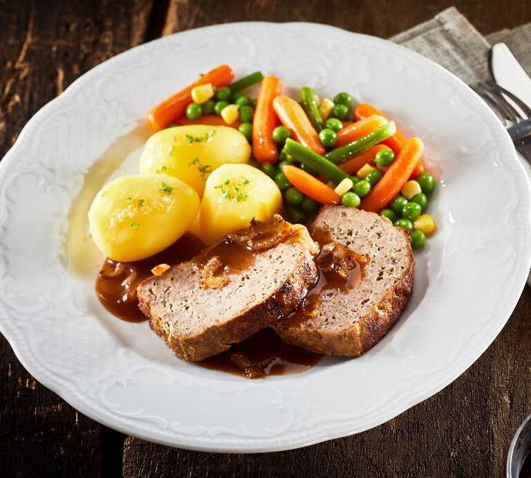 Draufsicht: Auf einem rustikalen Holztisch steht ein weißer Teller mit Omas Hackbraten mit Soße und Erbsen-Möhren-Bohnen-Gemüse. Daneben liegen eine Serviette aus grobem Leinen und ein Besteck. | © Adobe Stock / exclusive-design