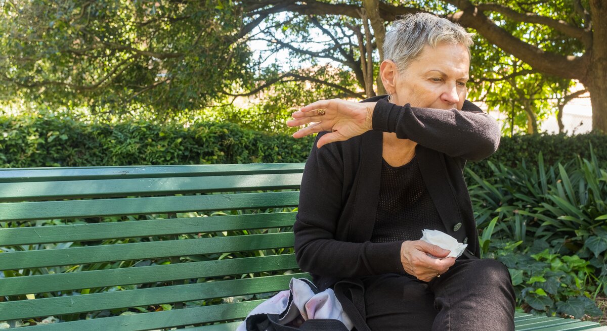 Eine ältere Frau mit kurzen grauen Haaren, die eine schwarze Jeans und ein schwarzes Oberteil trägt, sitzt auf einer grünen Parkbank im Freien. Sie hält ein Taschentuch in der rechten Hand und hustet in ihre Armbeuge.