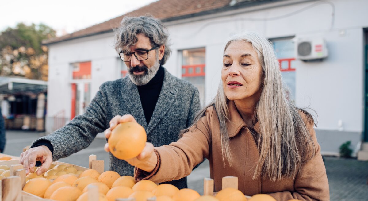  Ein älteres Paar mit grauen Haaren steht an einem Obststand und wählt gemeinsam frische Orangen aus. Der Mann, der eine Brille und einen grauen Wollmantel trägt, hält eine Orange hoch, während die Frau mit langen, glatten Haaren und einem braunen Mantel aufmerksam zusieht. Sie scheinen die Qualität der Früchte zu begutachten, was auf ein Bewusstsein für gesunde Ernährung hinweist. Im Hintergrund ist ein Teil eines Gebäudes mit einem roten Fensterrahmen zu sehen, was auf ein öffentliches Umfeld wie einen Markt hinweisen könnte.